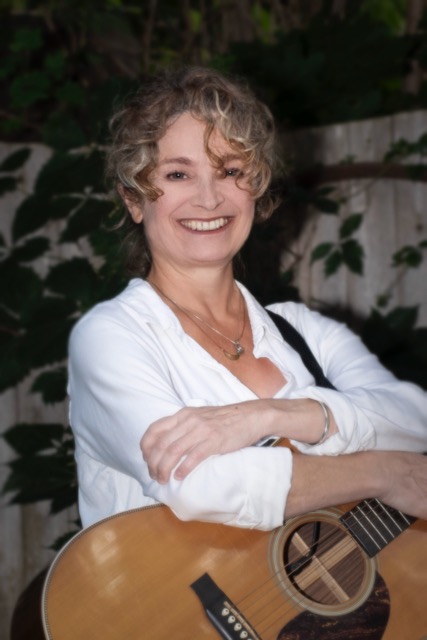 Patti Casey : white woman with light, curly hair smiling at camera. She is wearing a white shirt and her arms are resting on an acoustic guitar.