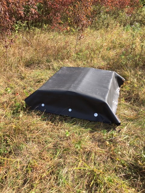 A black rubber material covers a rectangular object with a slight peak. The "snake hotel" sits in an autumn meadow with some woody shrubs visible in the background.